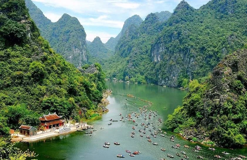 Khung cảnh Ninh Bình với dòng sông chảy quanh những ngọn núi hùng vĩ. Một ngôi chùa yên bình nằm dưới chân núi, trong khi các con thuyền nối đuôi nhau chèo đến viếng thăm trong lễ 