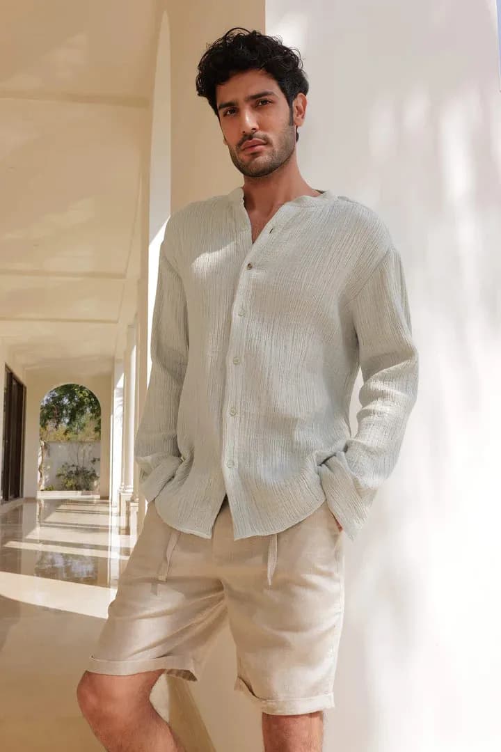 A young man wearing white clothes stands outdoors on a sunny day, with the bright sunlight highlighting his casual and relaxed look against a clear sky