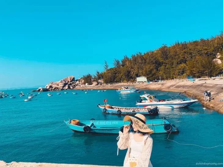 Woman by Quy Nhon’s serene blue beach, boats docked nearby, with a tranquil azure sky overhead.