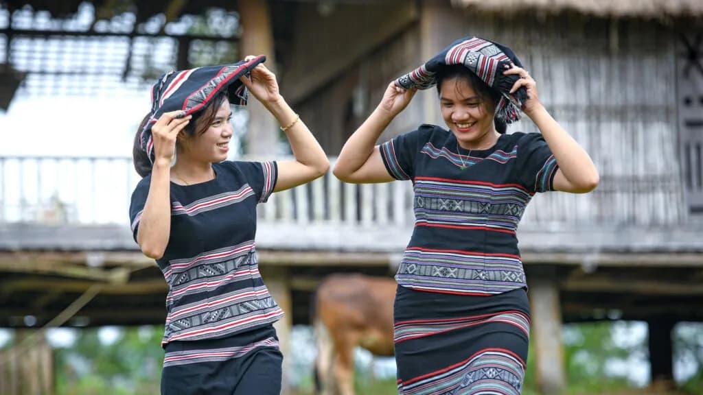 Two Hrê girls smiling brightly in the sun, wearing traditional woven scarves.